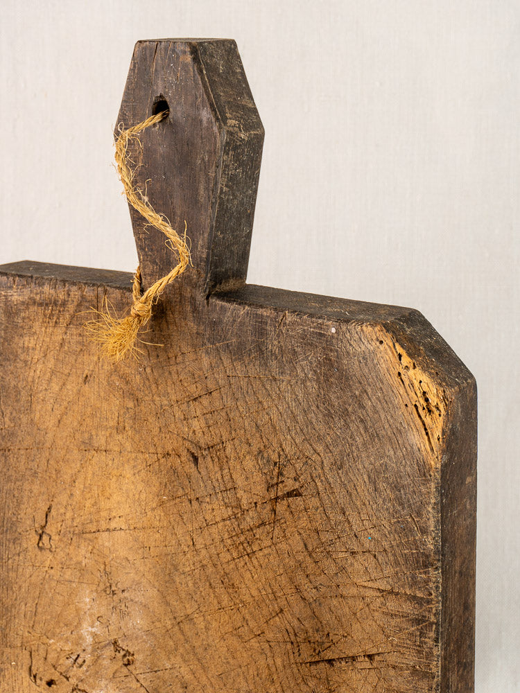 Antique French cutting board, hand carved with diamond handle, worn surface showing knife marks and frayed twine threaded through handle.