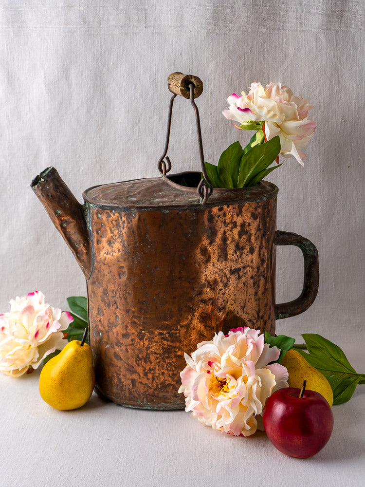 19th-century French hammered copper water carrier with flowers, yellow pear, and red apple on light background.