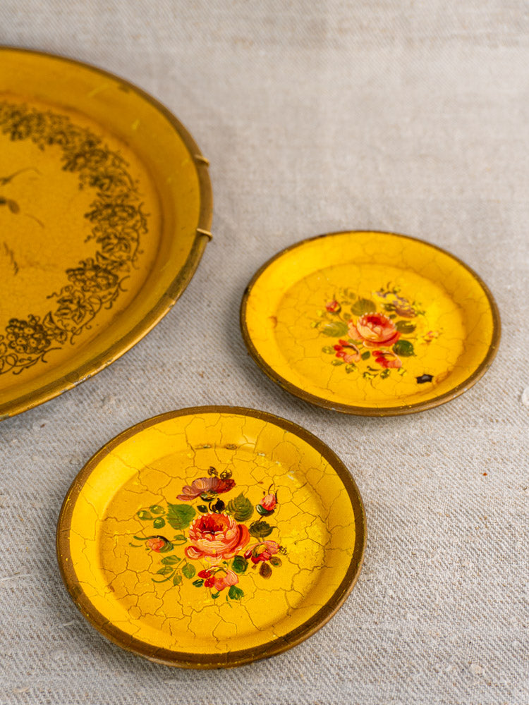 Antique French yellow lacquered tôle tray set with small floral plates, visible cracks, textured fabric background.
