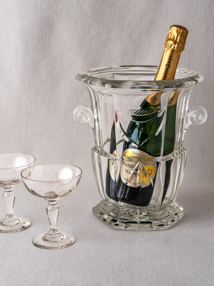 French cut crystal champagne bucket with bottle, beside two vintage-style coupes on a light surface.