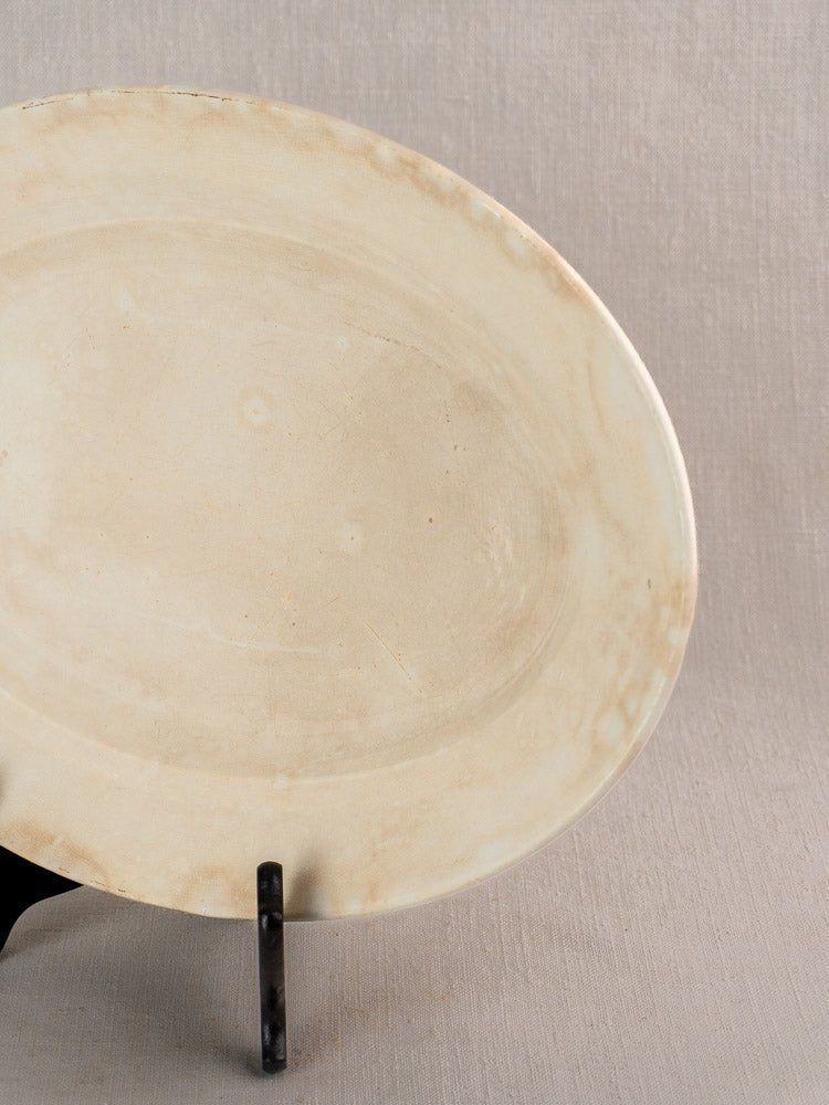 Oval vintage French Sarreguemines ivory ironstone platter with discoloration, upright on black stand, beige backdrop.