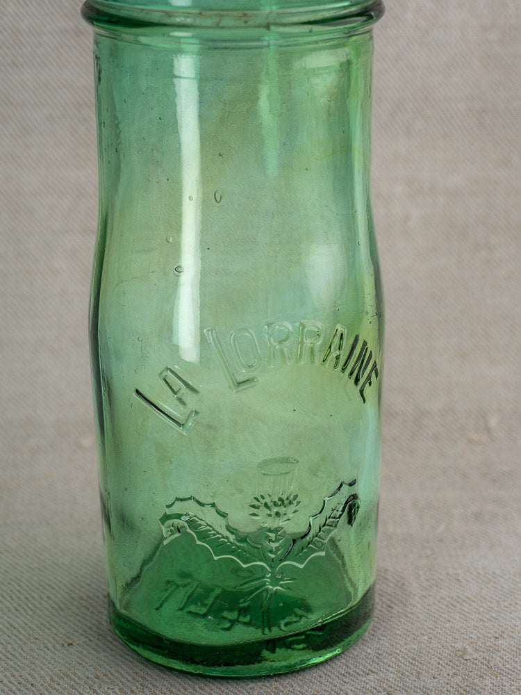 Pair of antique French La Lorraine green glass jars with thistle motifs on textured beige surface.