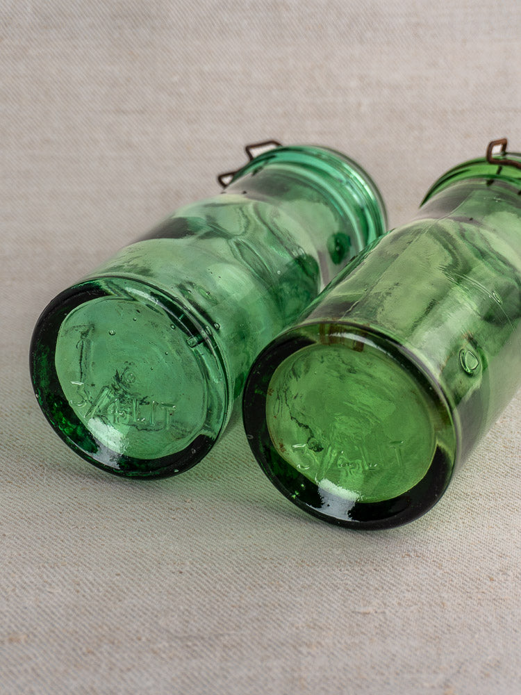 Pair of antique French La Lorraine green glass jars with thistle motif, embossed details, and metal clasp lids on neutral fabric.