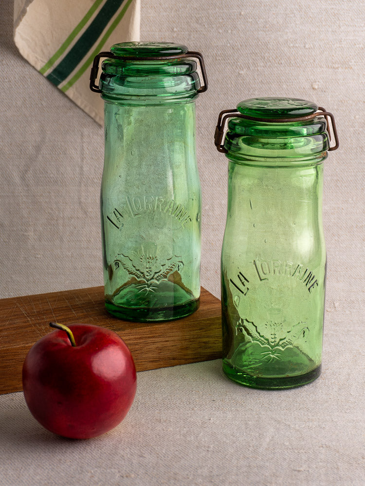 Pair of antique French La Lorraine green glass jars with thistle motif on neutral fabric background.