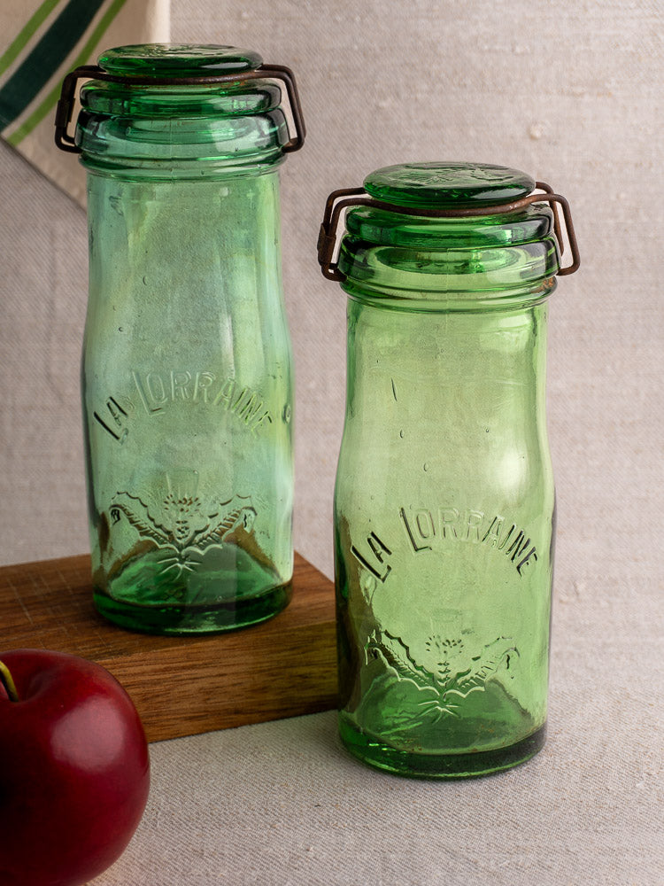 Pair of antique French La Lorraine green glass jars with thistle motif, on beige surface near red apple and striped cloth.