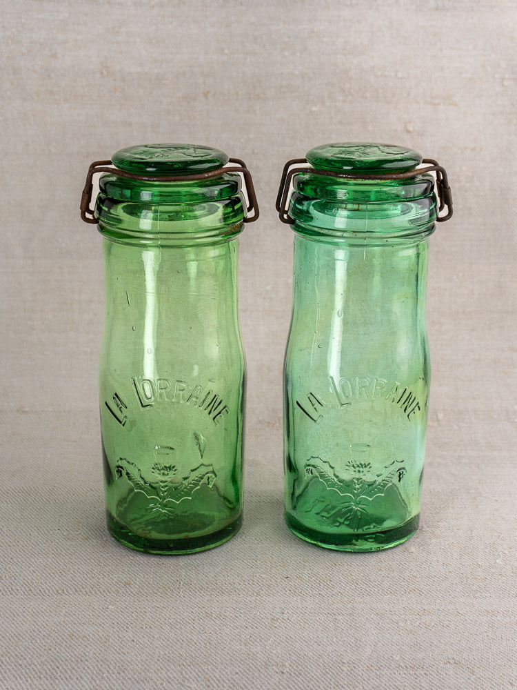 Pair of antique French La Lorraine green glass jars with thistle motif on neutral fabric background.