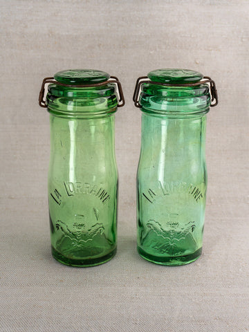 Pair of antique French La Lorraine green glass jars with thistle motif on neutral fabric background.