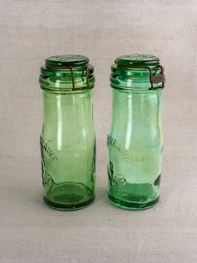 Antique French La Lorraine green glass jars with embossed thistle motifs and metal closures on neutral fabric.