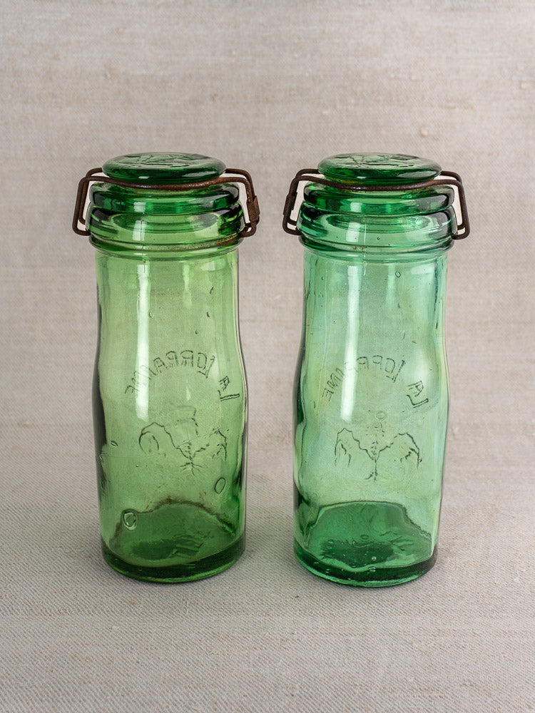 Pair of antique French La Lorraine green glass jars with thistle motif and metal clasps on textured fabric.