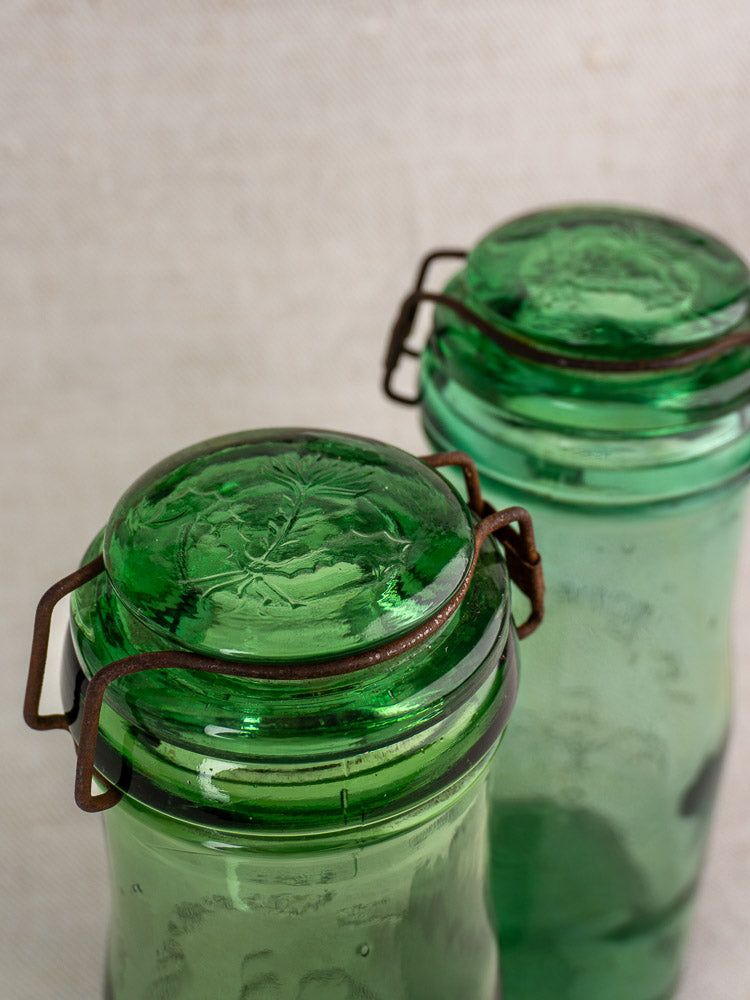 Antique French La Lorraine green glass jars with thistle lids and metal clasps on a textured neutral backdrop.