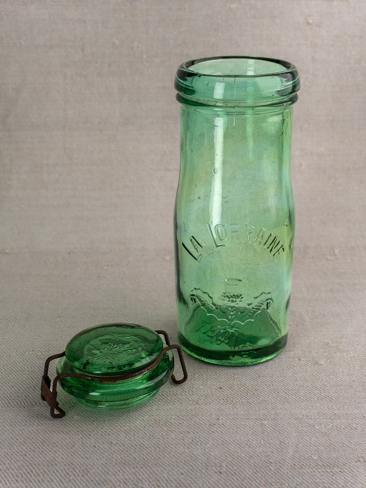 Pair of antique French La Lorraine green glass jars with thistle motifs, embossed fronts, and metal-clasp lids on beige fabric.