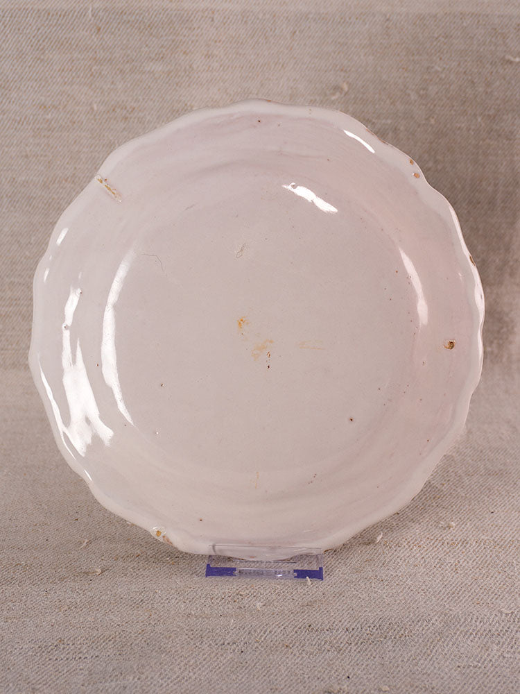 Antique French faience plate with hand-painted turnip motif, off-white glaze, scalloped edge, on stand against beige background.
