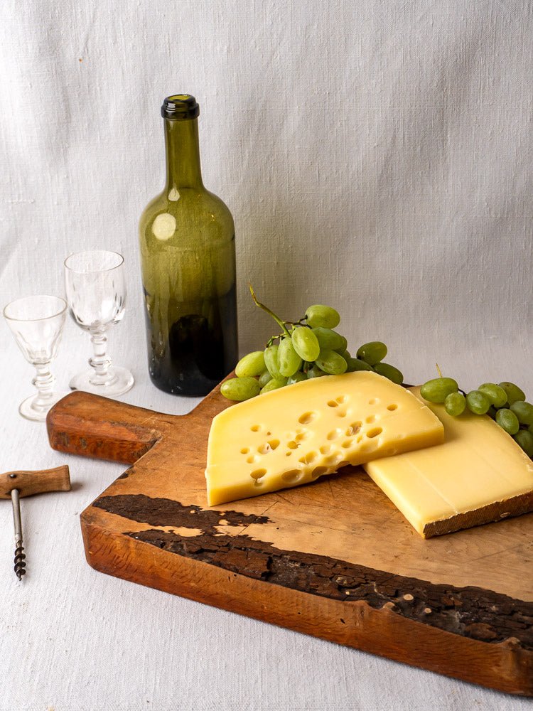 Antique 19th-century French live edge cutting board with cheese, grapes, wine bottle, and glasses on white linen.