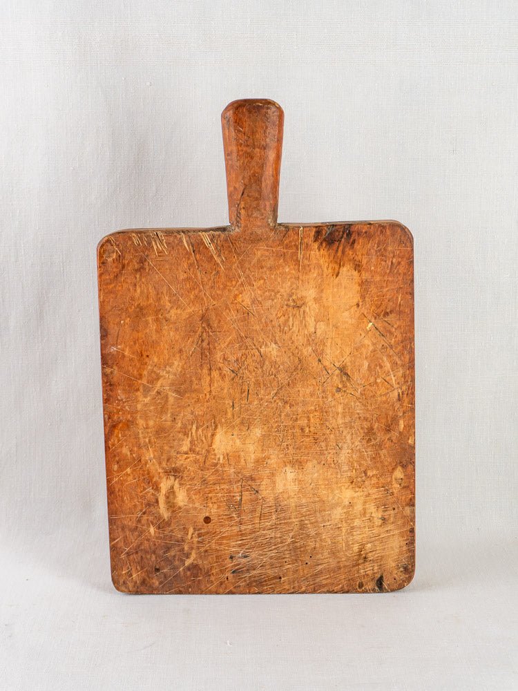 Antique 19th-century French live edge cutting board with short handle, upright on light gray background.