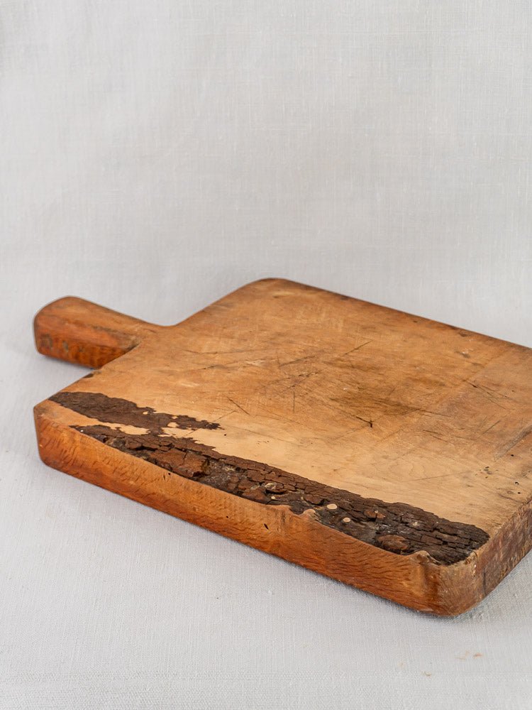 Antique French live edge cutting board with handle and aged, textured surface on light gray fabric.