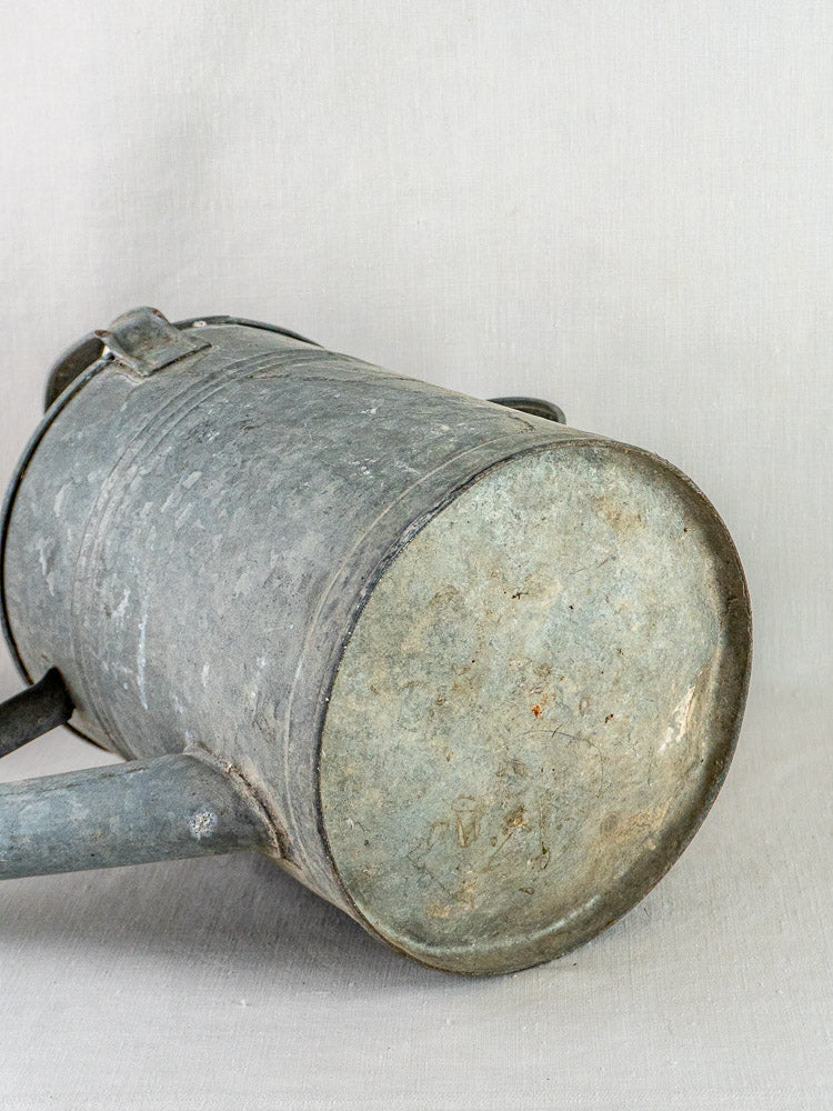 French galvanized zinc long-spout watering can resting sideways, round base and spout visible on neutral surface.