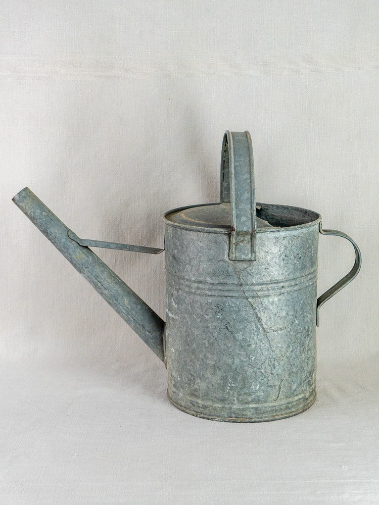 French galvanized zinc watering can with curved handle and long spout on a light fabric background.