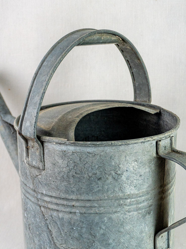 French galvanized zinc long-spout watering can with curved handle and weathered finish on plain background.