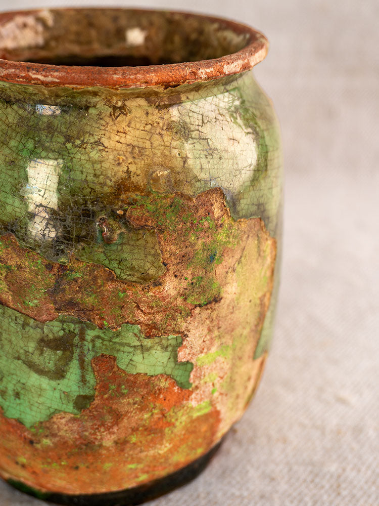 Antique French confit pot with green glaze, cracked surface, chipped edges, and red-brown patina on neutral background.
