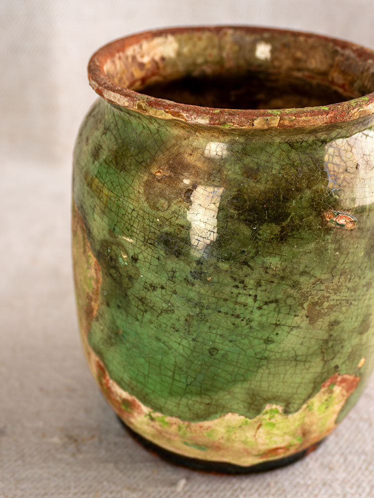 Antique French confit pot with green glaze, crackled finish, and uneven rim on neutral fabric background.