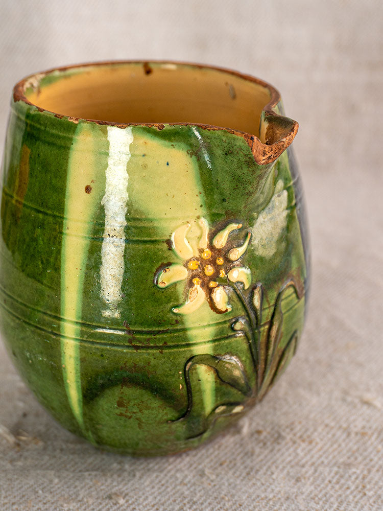 Antique French Jaspe Savoie earthenware pitcher with green glaze, yellow-white floral motif, chipped spout, on fabric background.