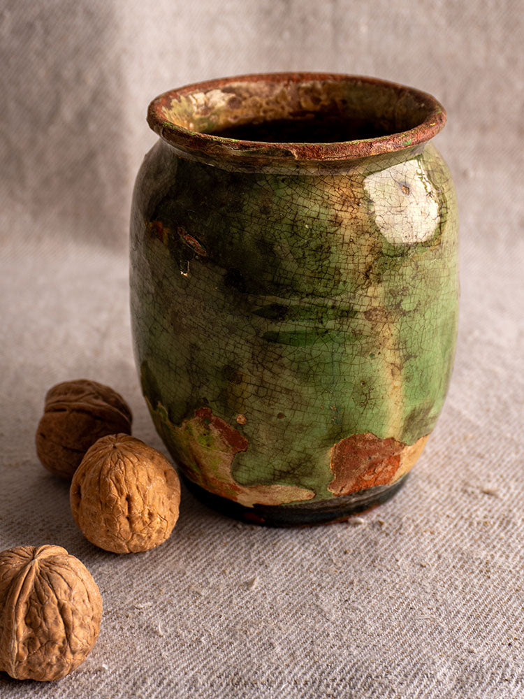 Antique French confit pot with green glaze and weathered ceramic on textured fabric beside three walnuts.
