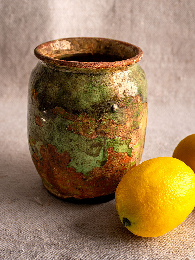Antique French confit pot with weathered green glaze beside two lemons on a textured beige surface.