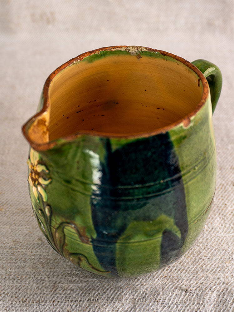 Antique French Jaspe pottery pitcher in Savoie earthenware with green glaze and floral motif, on a textured neutral surface.