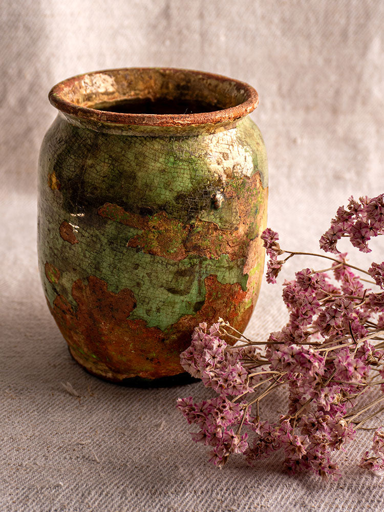 19th-century French confit pot with green glaze displayed on beige fabric next to dried pink flowers.