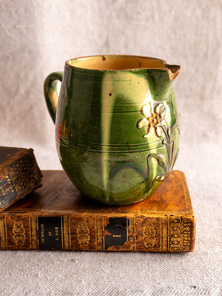Antique French Jaspe pottery pitcher with green glaze and floral motif on vintage books, beige fabric background.