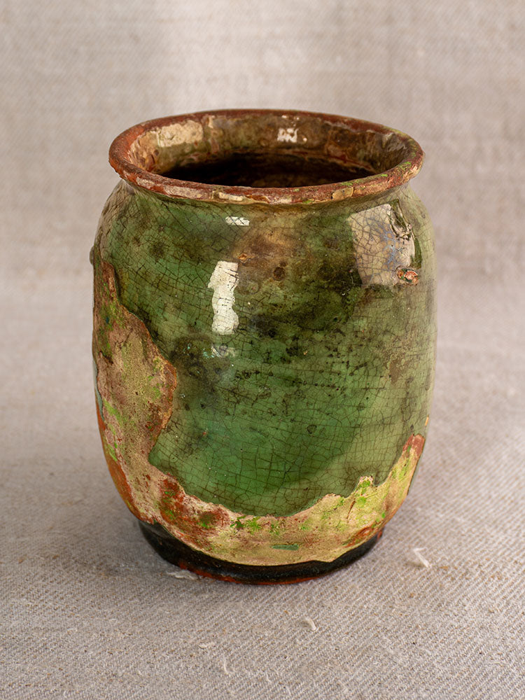 Antique French ceramic confit pot with green and brown mottled glaze, weathered finish, on light gray textured fabric.