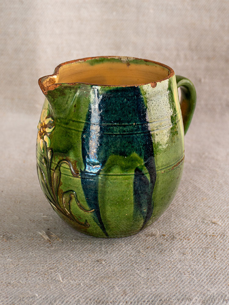 Antique French Jaspe pottery pitcher with green glaze, floral design, spout, handle, and chipped rim on beige textured fabric.