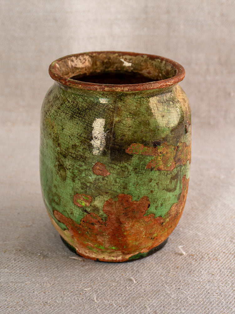 19th-century French confit pot with cracked surface, mottled green glaze, and reddish-brown ceramic on textured beige fabric.