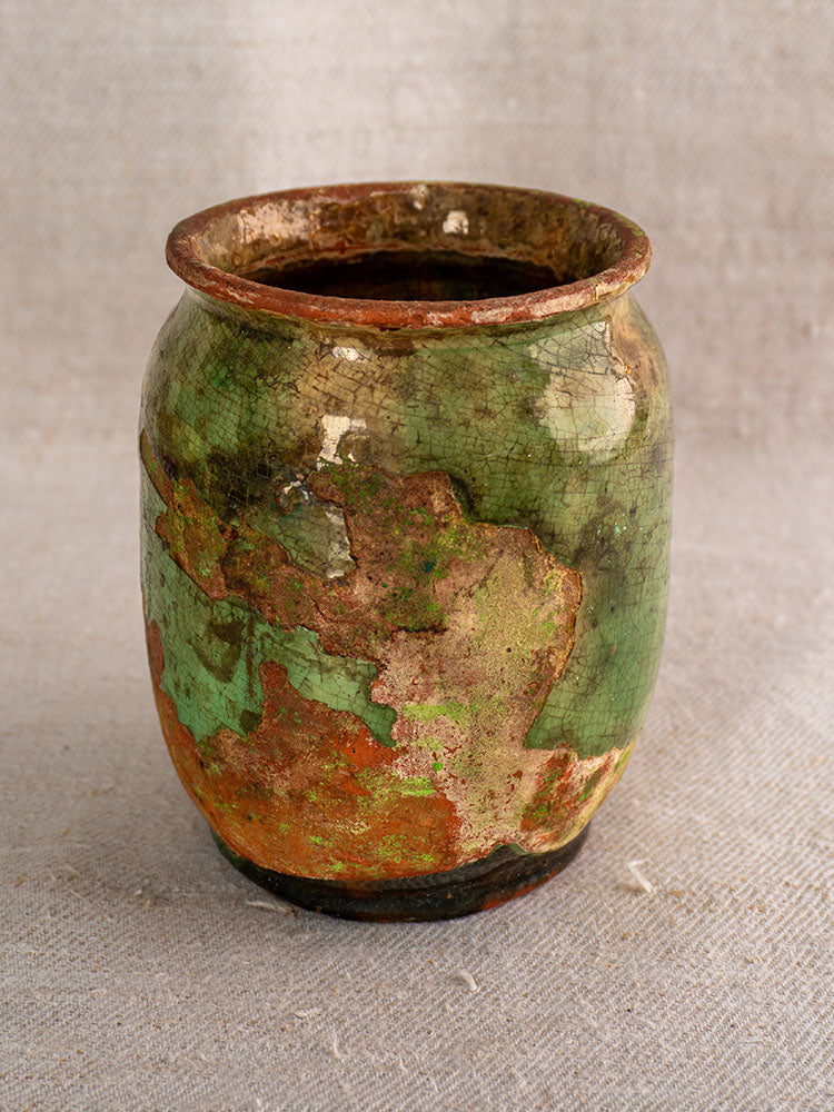 Antique French confit pot with weathered green and brown glaze on a textured beige fabric background.