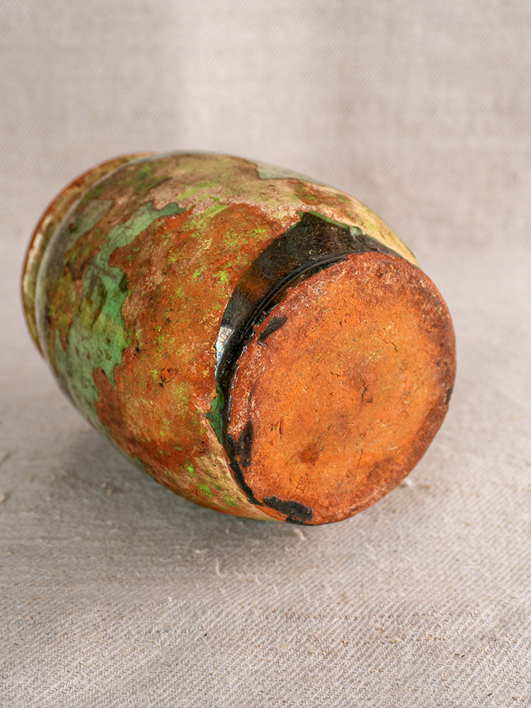 19th-century French confit pot with green glaze, rusted base, and weathered green-brown markings on light textured fabric.