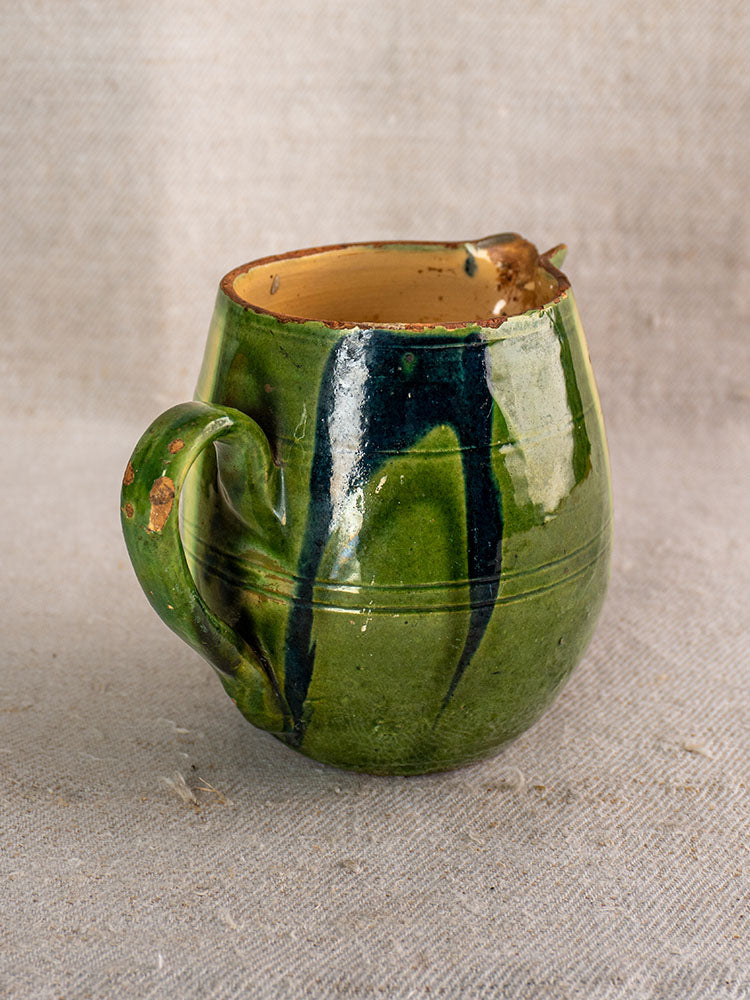 Antique French Jaspe pottery pitcher with green glaze and floral design, brown accents, on textured beige fabric.