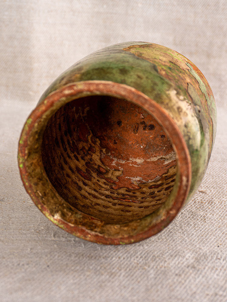 19th-century French confit pot with green glaze, displayed on neutral textured fabric, showing aged exterior and rustic interior.