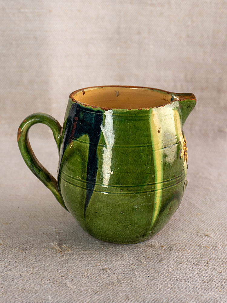 Antique French Jaspe pottery pitcher in green-glazed Savoie earthenware with florals on a textured beige fabric backdrop.