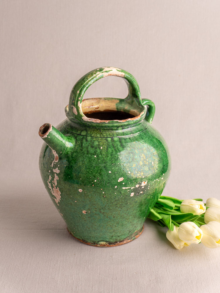 Antique French Provençal green glazed earthenware gargoulette with large handle, some chips, beside white tulips on light surface.
