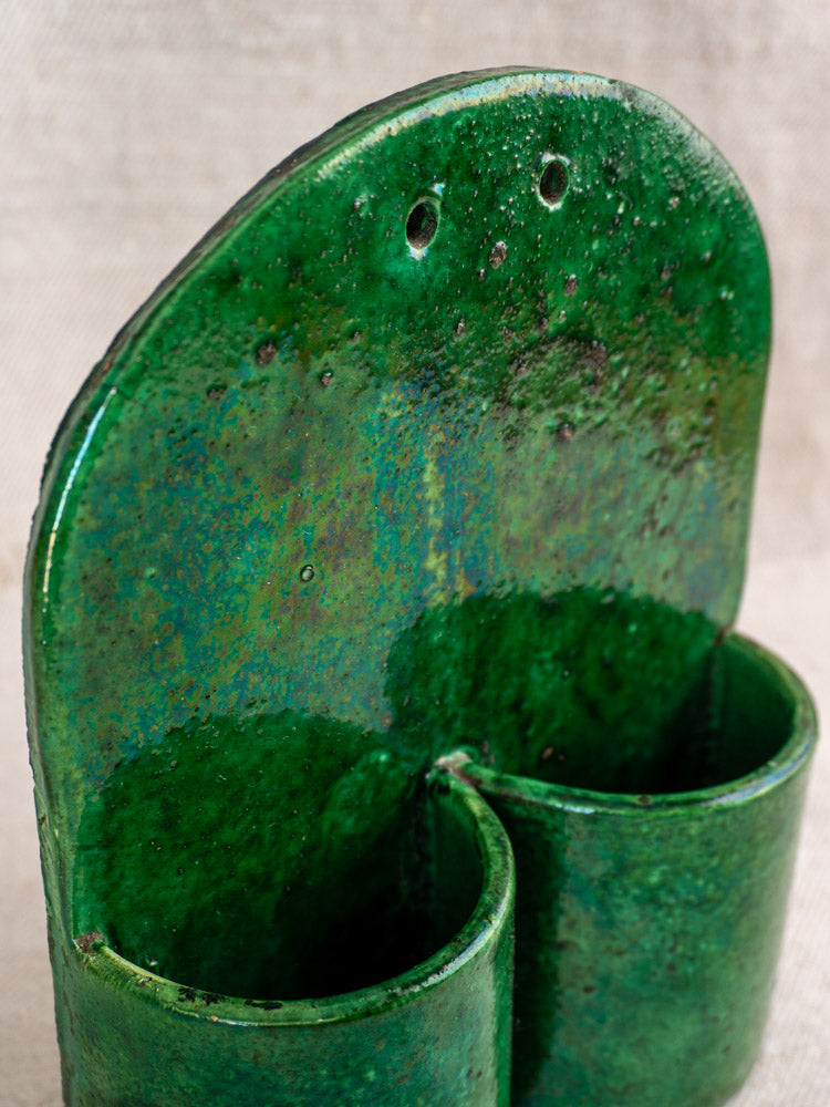 Antique French green glazed earthenware wall utensil holder with two compartments, set against textured fabric.