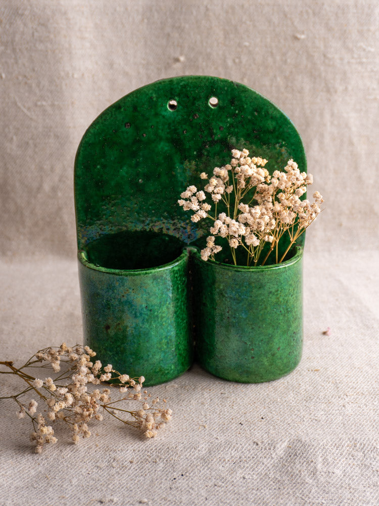 Antique French green glazed earthenware wall utensil holder with dried white flowers, on beige textured fabric.