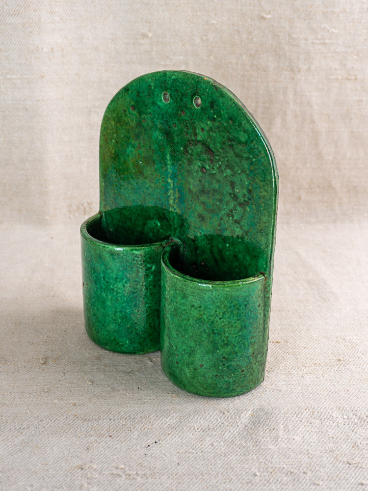 Antique French wall utensil holder with two green glazed earthenware cylinders on curved backing, beige fabric background.