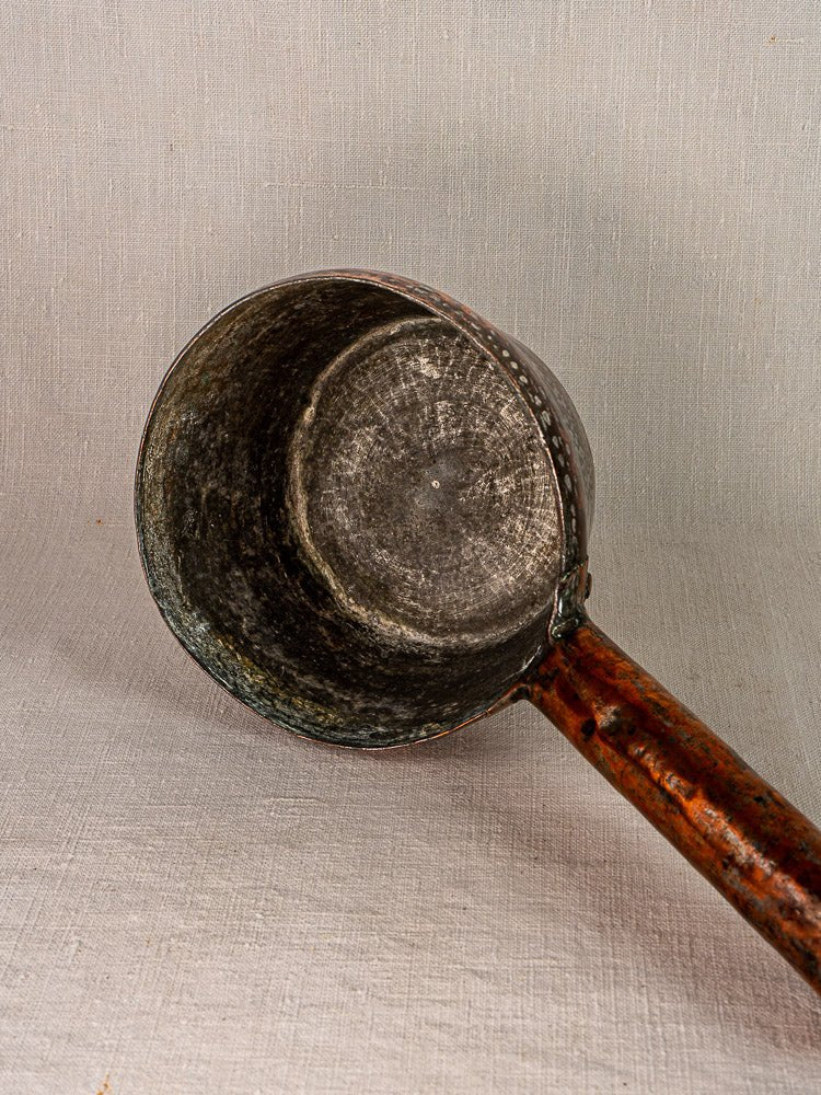 Vintage French hand-forged copper saucepan with deep round bowl and long wooden handle on light textured surface.