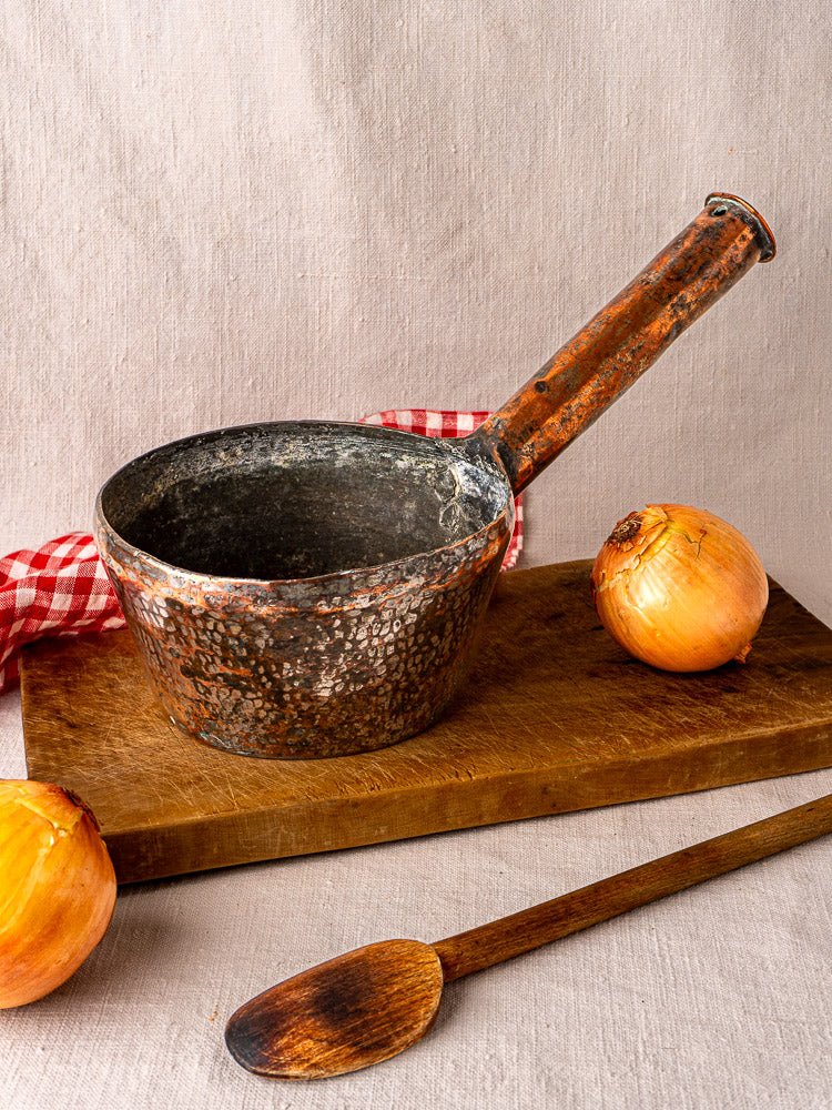 Antique French copper saucepan with wood handle on cutting board, onions, spoon, checkered cloth.