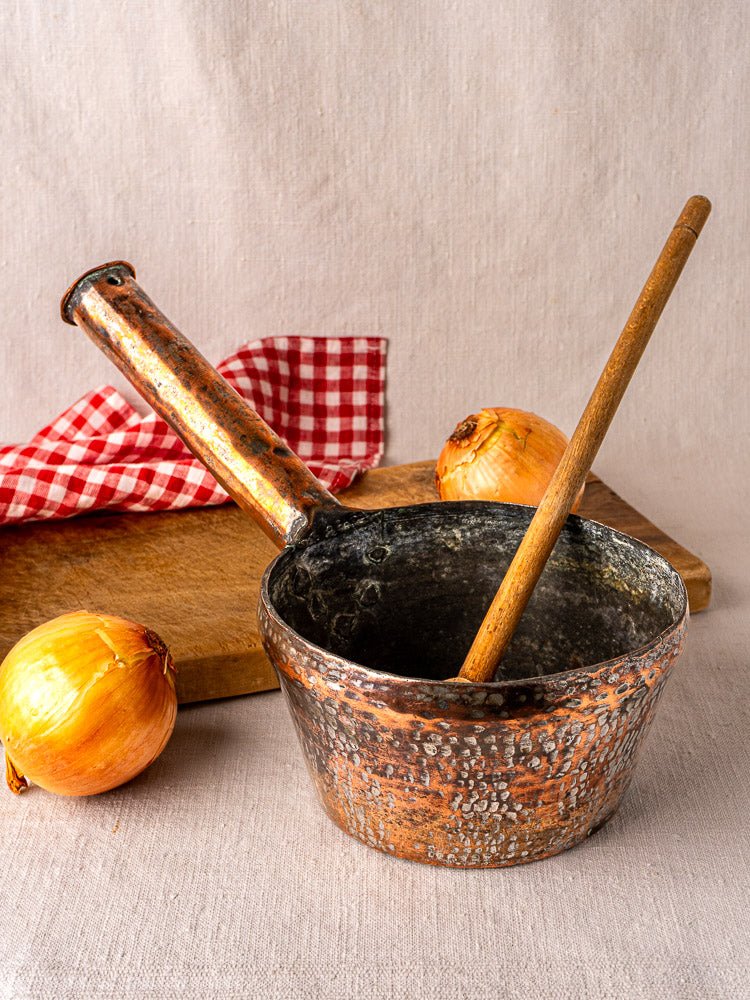 Antique copper pot with wooden spoon on a wooden cutting board with onions and a checkered cloth.