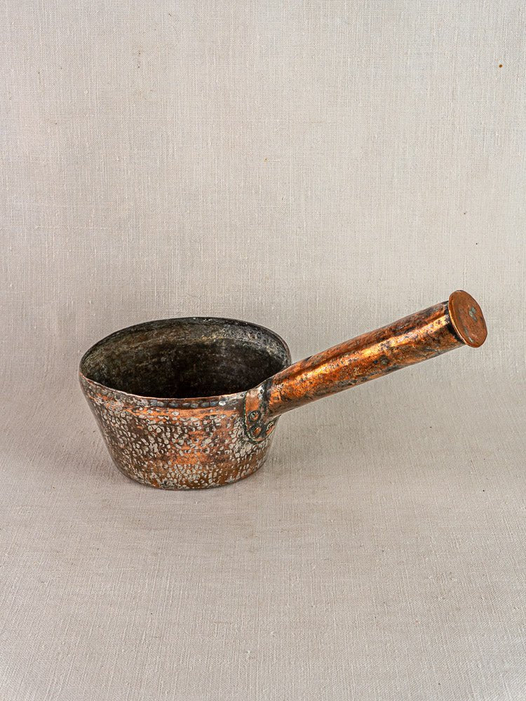 Antique French hand-forged copper saucepan with patina and long angled handle on light fabric.
