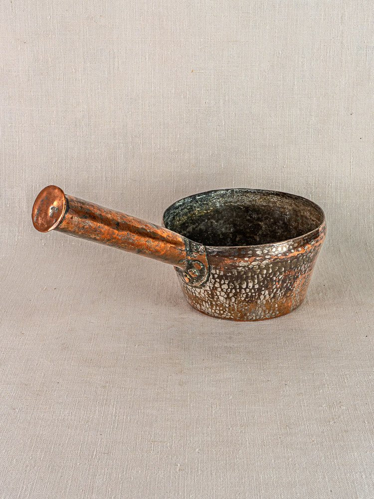 Antique French hand-forged copper saucepan with hammered finish and long handle on neutral fabric.