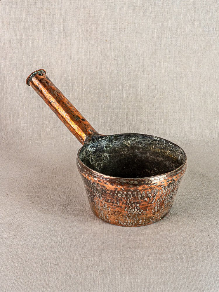 Antique French hand-forged copper saucepan with long handle, vintage patina and wear, on fabric.