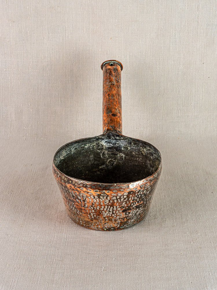 French antique copper casserole à queue with long handle, speckled finish on beige fabric background.