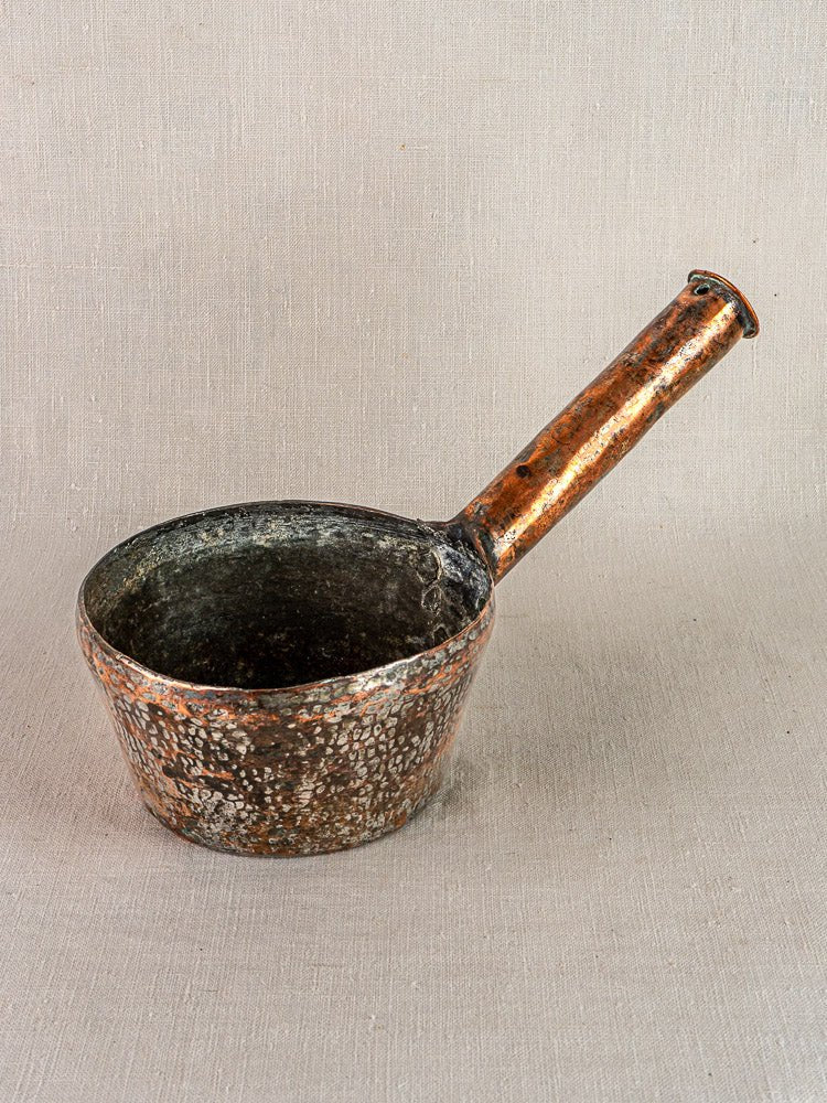 Vintage French hand-forged copper saucepan with deep bowl and long handle on beige textured surface.